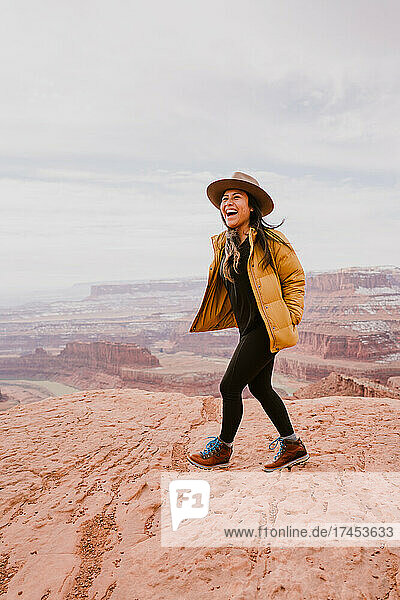 A woman wearing brimmed hat laughs while hiking above desert canyon