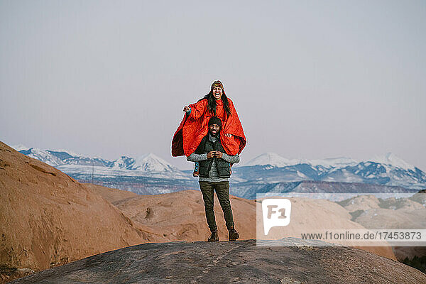 A woman sits on her friend's shoulders while laughing in the mountains