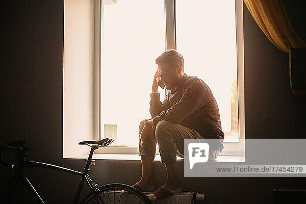 Man talking on smart phone sitting on windowsill at home