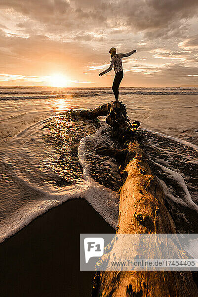 Teen girl balancing on log at a beach in New Zealand at sunrise