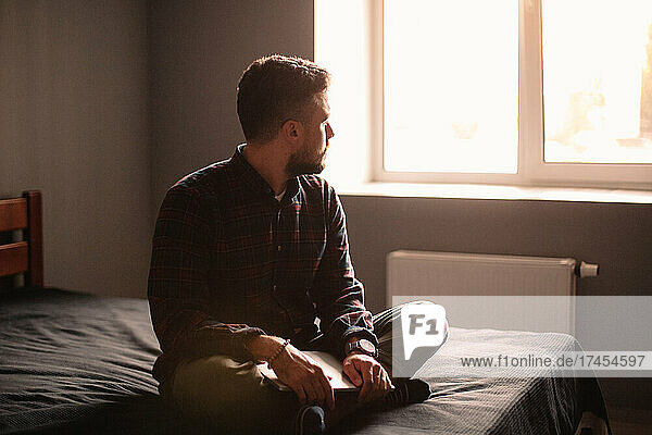 Thoughtful man with laptop looking through window sitting on bed