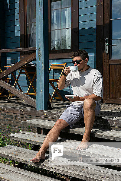 A young man drinks coffee on the porch of the house
