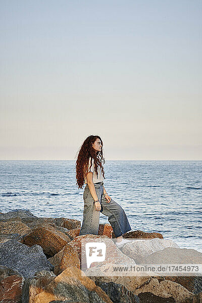 Young redhead woman looks out to sea while she is standing on rocks