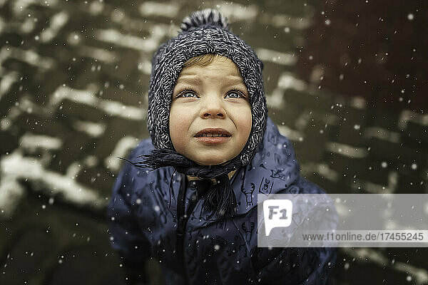 Small boy in blue winter clothes looking up at the snowing sky
