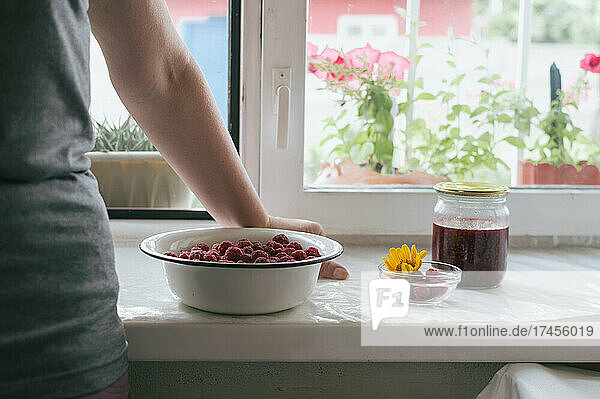 Woman leans on windowsill with bowl of raspberries and jam jar