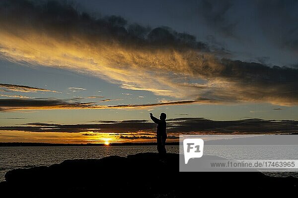 Frau fotografiert spektakulären Sonnenuntergang mit Handy auf einem Felsen stehen an der Küste des atlantischen Ozeans vor Concarneau  Département Finistère  Bretagne  Frankreich  Europa