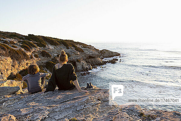 Teenage girl and young brother at sunset  sitting side by side looking over the ocean.