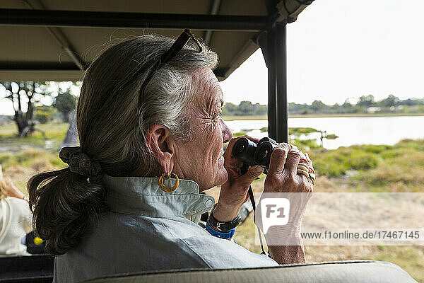 Senior woman using binoculars  sitting in a safari vehicle  looking out over marshes and waterway