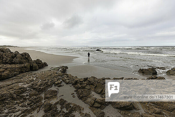 A man walking across sand to the water's edge on a beach  overcast day and surf waves breaking on shore.