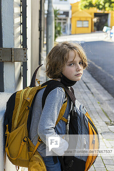 A boy with a black facemask tucked under his chin  on a street with a backpack and bag.
