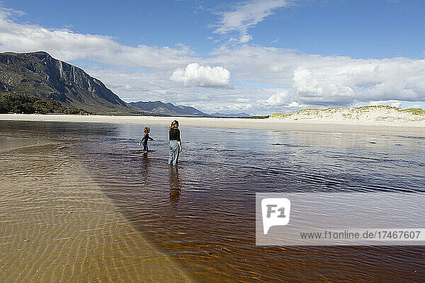 Teenage girl and young boy on an open sandy beach wading through shallow water.