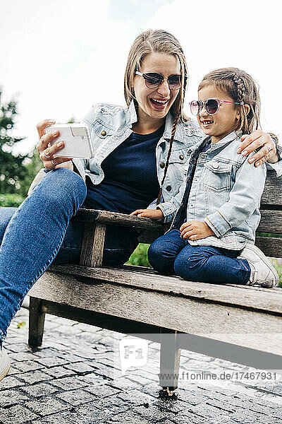 Happy woman sharing smart phone with daughter while sitting on bench in park