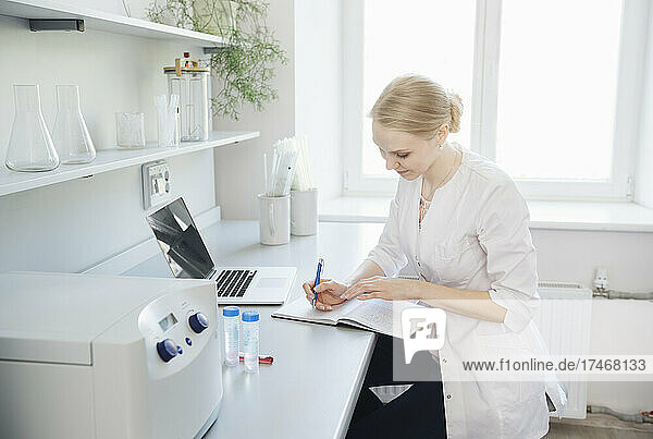 Blond scientist with laptop writing in note pad at desk