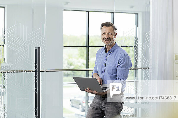 Smiling businessman with laptop leaning on glass door