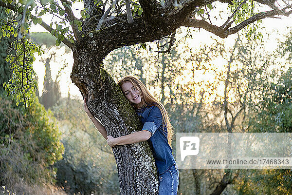 Lächelnde junge Frau umarmt einen Baum auf dem Land