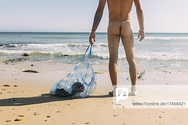 Naked volunteer collecting plastic pollution in mesh bag at beach