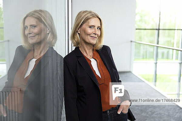 Smiling businesswoman with hand in pocket leaning on glass at corridor