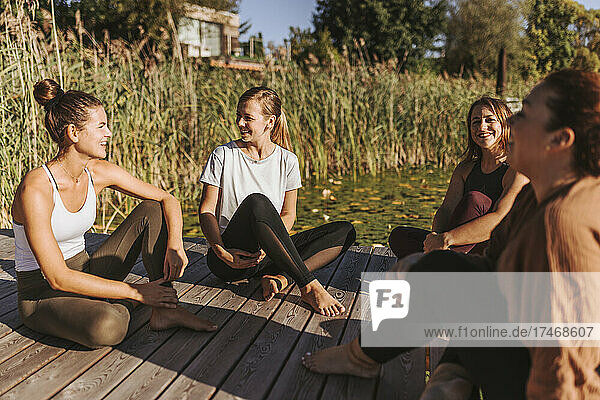 Female friends talking to each other while sitting on jetty by lake
