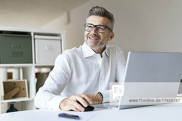 Smiling businessman with laptop contemplating in office