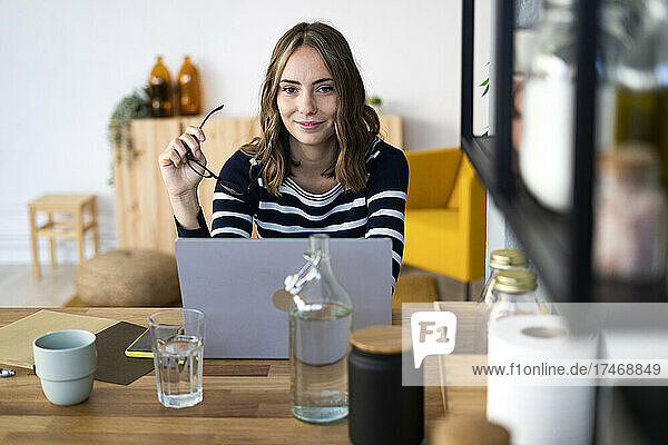 Smiling woman holding eyeglasses in front of laptop at kitchen