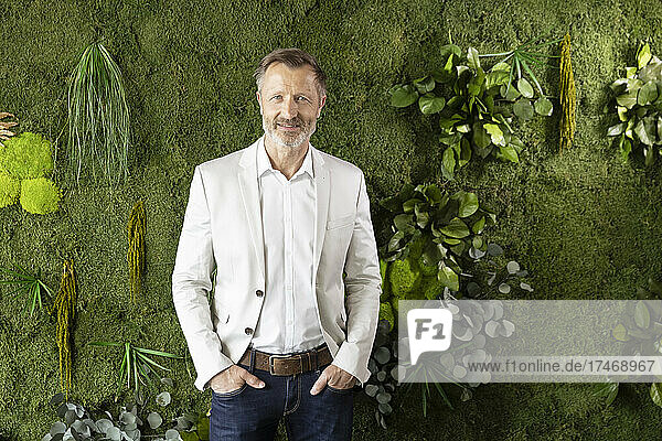 Mature businessman standing with hands in pockets by plants