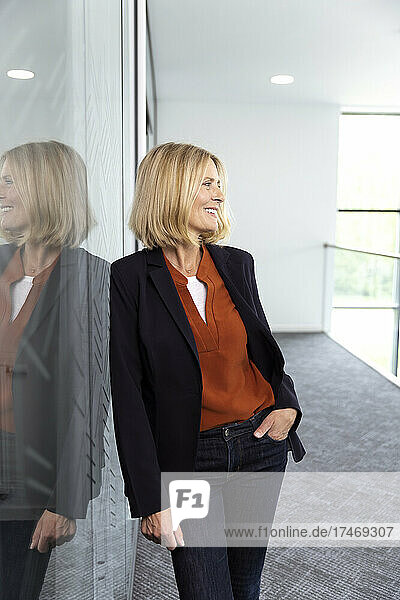 Smiling businesswoman with hand in pocket leaning on glass
