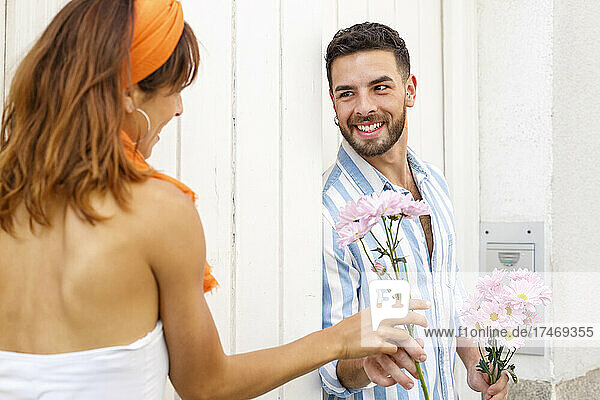 Smiling man giving bunch of flowers to woman leaning on wall