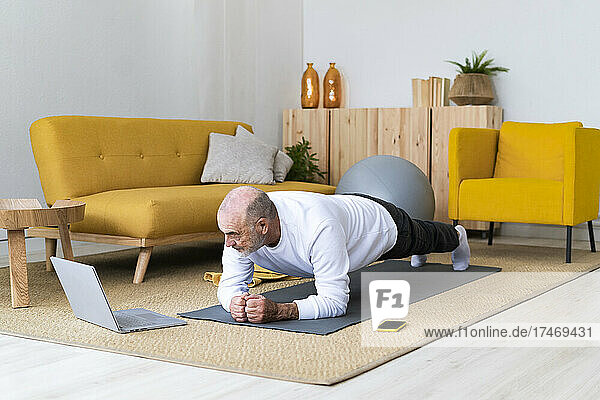 Senior man with laptop practicing plank pose on exercise mat