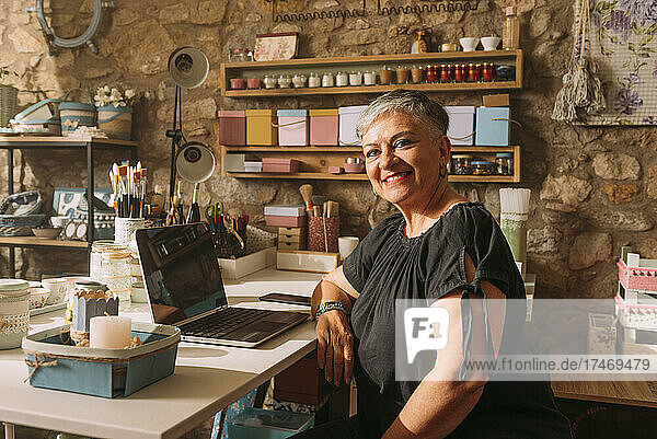 Smiling craftswoman with laptop sitting in workshop