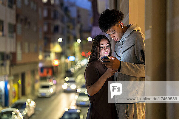 Multi-ethnic couple using smart phone while standing on balcony at night