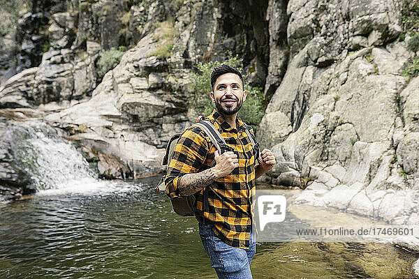 Smiling mid adult man wearing backpack standing by pond in forest