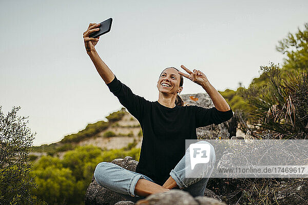 Smiling woman gesturing while taking selfie through smart phone on rock