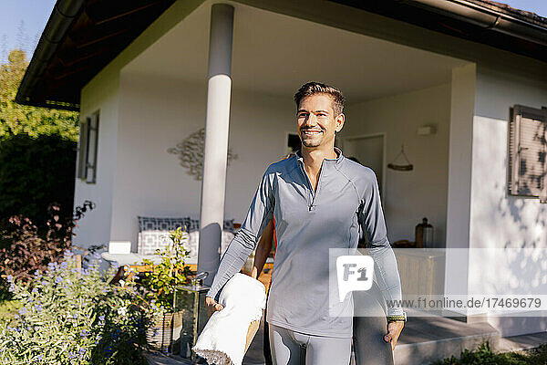 Smiling man walking with exercise mat in front of house