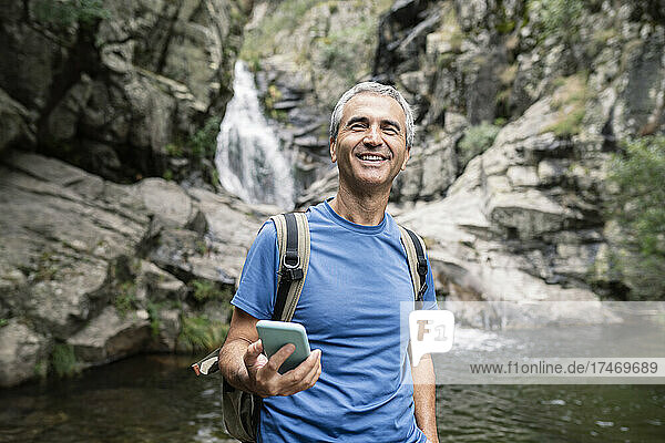Smiling male tourist holding mobile phone in forest