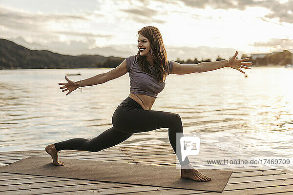 Smiling woman with arms outstretched doing yoga on jetty during sunset