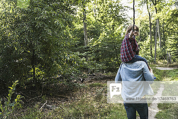 Junge mit Stock sitzt auf den Schultern eines Mannes im Wald