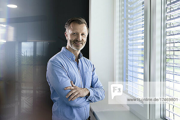 Smiling mature businessman standing with arms crossed by window