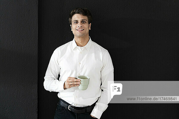 Selbstbewusster Geschäftsmann mit Kaffeetasse steht vor schwarzer Wand