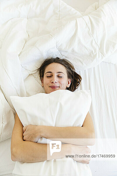 Smiling young woman embracing pillow while sleeping on bed