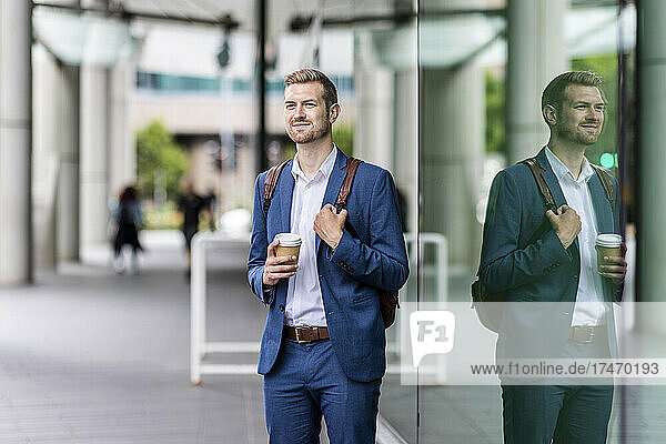 Male professional with backpack and disposable cup standing by glass wall