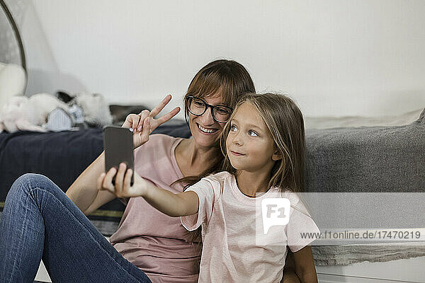 Girl taking selfie with mother showing peace sign through mobile phone at home