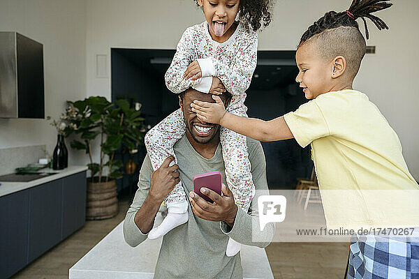 Junge bedeckt die Augen seines Vaters  der sein Mobiltelefon in der Hand hält und seine Schwester auf der Schulter trägt