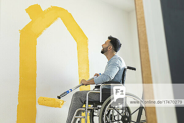 Smiling man holding paint roller while sitting on wheelchair in living room