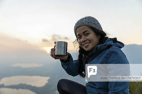 Smiling woman showing coffee mug on sunset during vacation