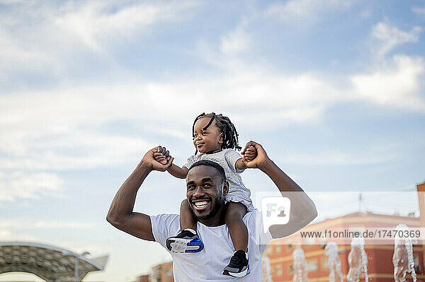 Happy man carrying daughter on shoulders under sky