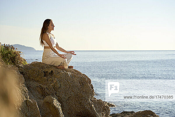 Sportswoman meditating in lotus position on rock at sunset