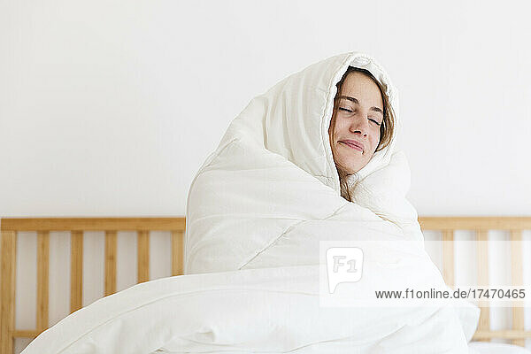 Lächelnde junge Frau mit geschlossenen Augen  zu Hause in eine weiße Decke gehüllt