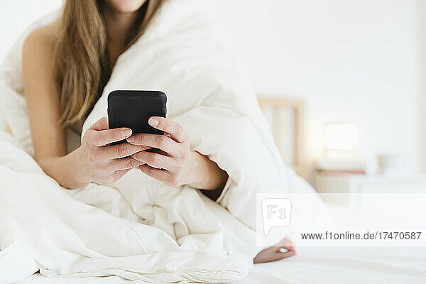 Junge Frau benutzt Mobiltelefon  während sie im Schlafzimmer auf dem Bett sitzt