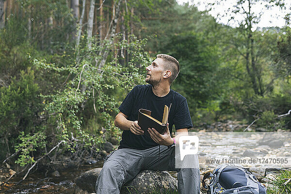 Man with diary sitting on rock in forest