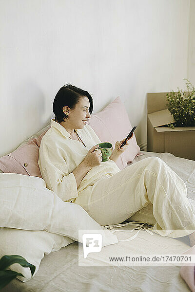 Mature woman with coffee cup using smart phone in bedroom